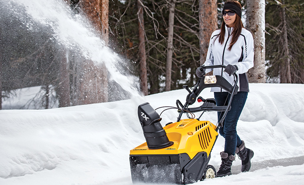 A person in winter gear and eye protection using a yellow snow blower.