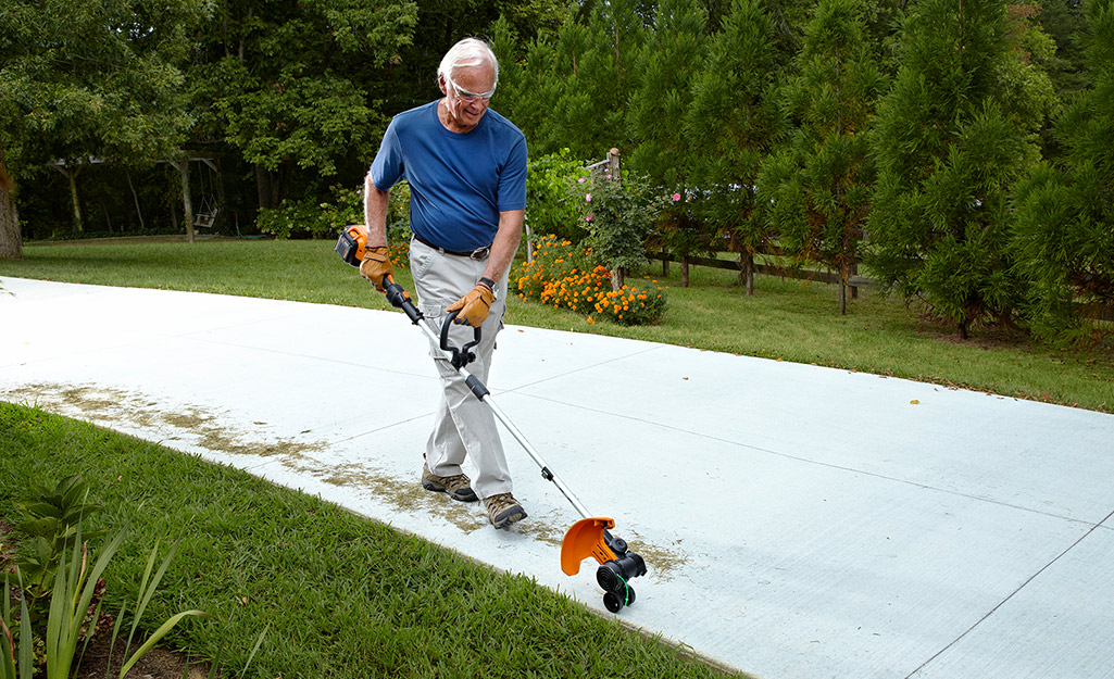 A man uses a string trimmer to edge the lawn by his driveway.