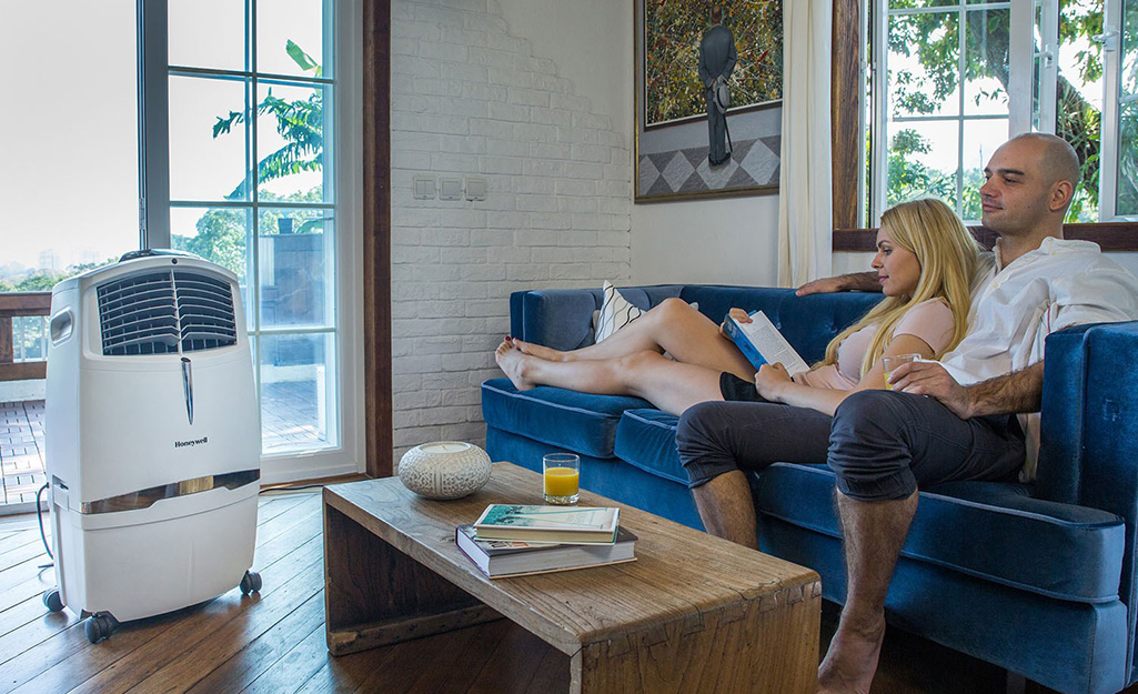 A couple sits on a blue couch next to a coffee table and an evaporative cooler. 