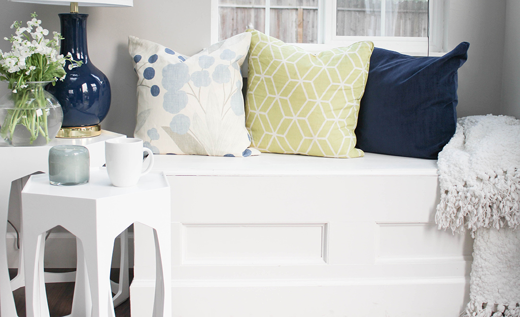 Three large throw pillows sit on top of a white bench next to white side tables holding a lamp, flowers and a coffee cup.