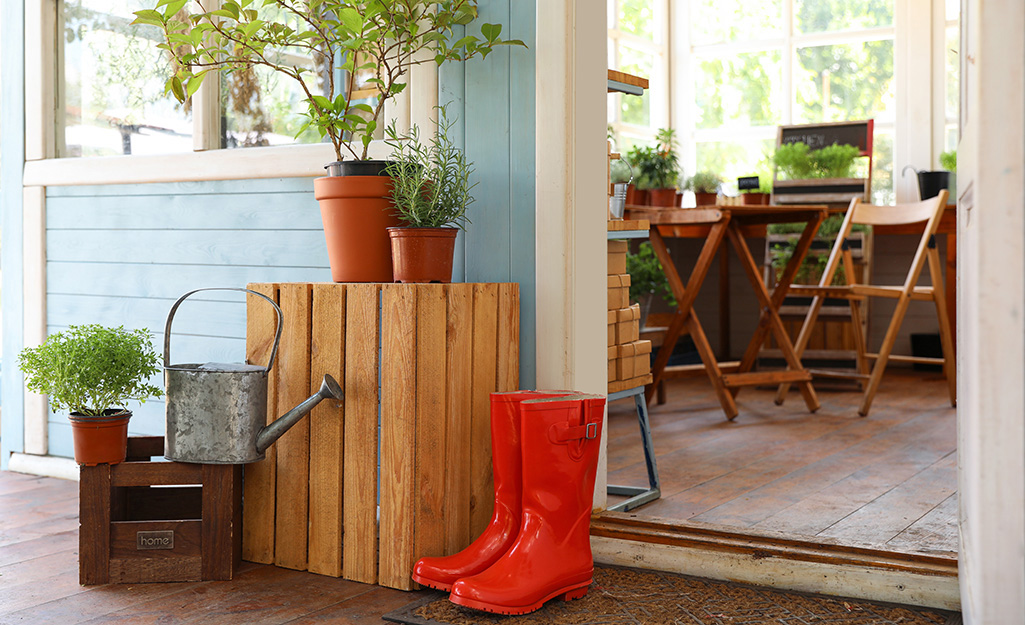 Rain boots, a watering can and wooden crates with potted plants on top stand by the open door of a garden-themed she shed.