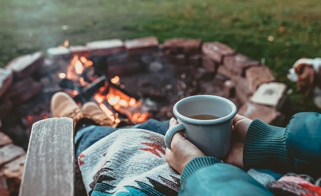 A person sitting beside a fire pit, holding a drink. A person sitting beside a fire pit, holding a drink.