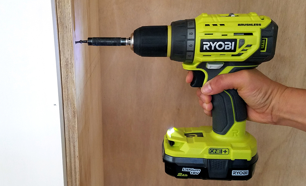 A person using a power drill to drill into a piece of wood against a wall.