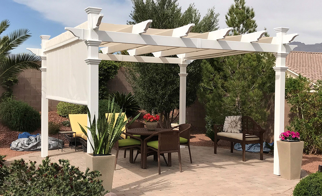 A white pergola stands above an outdoor table and next to plants in containers and shrubs