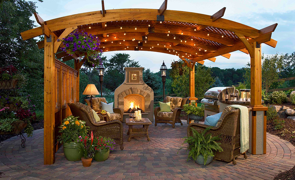 A pergola stands above a patio with wicker chairs, an outdoor fireplace and a grill.