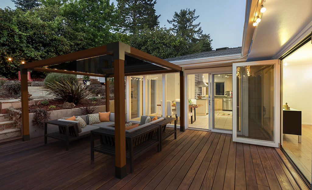A pergola stands above two outdoor couches on a deck in front of several open glass doors to a house.