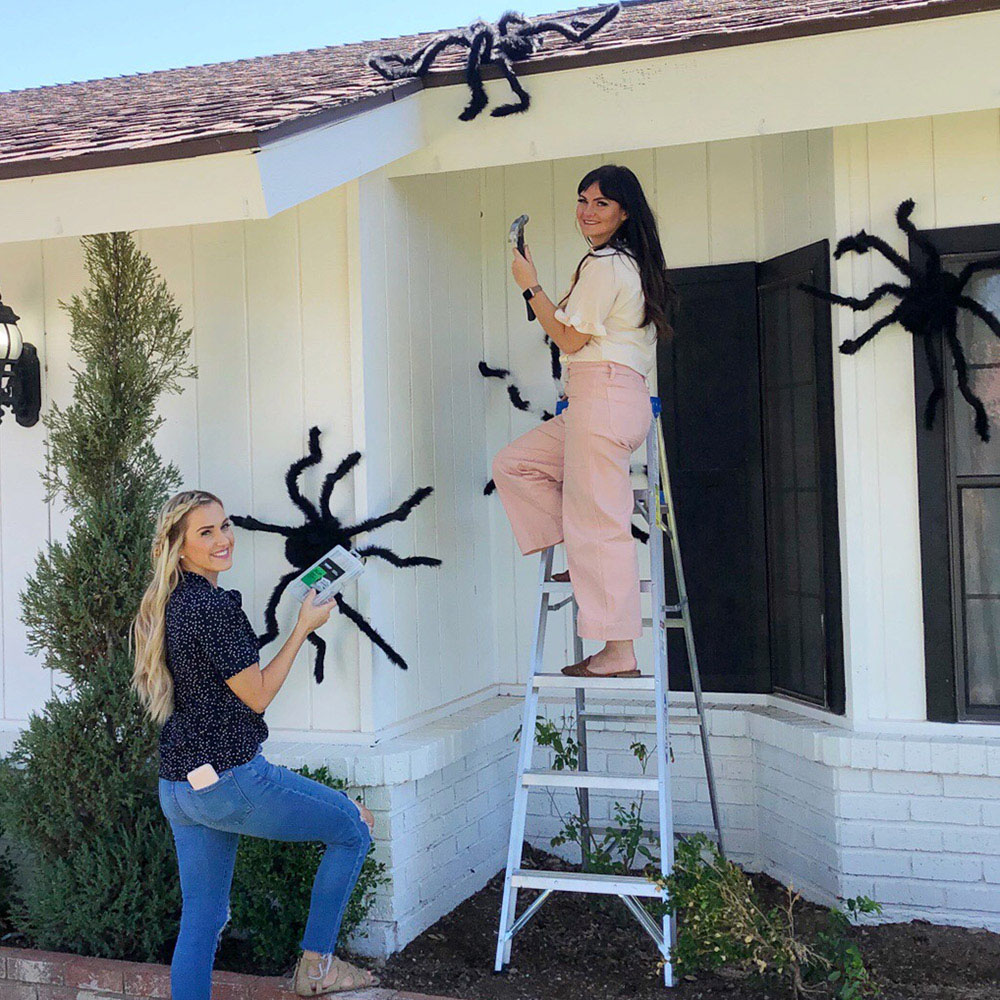 Two women standing in front of a white house decorated with black spiders for Halloween.
