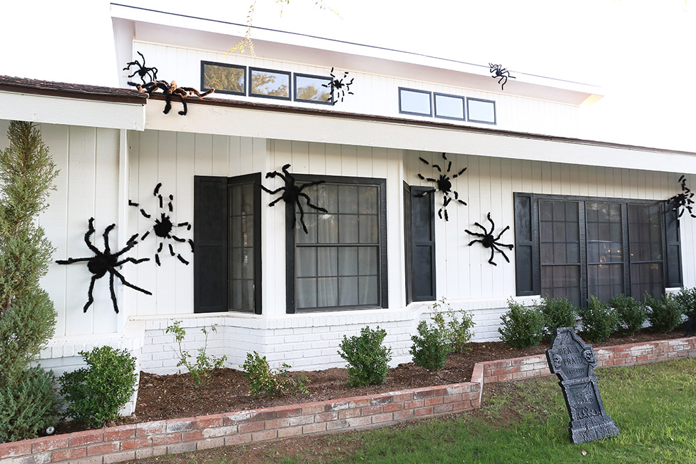 A tombstone in yard sits in front of a home covered with large spiders.