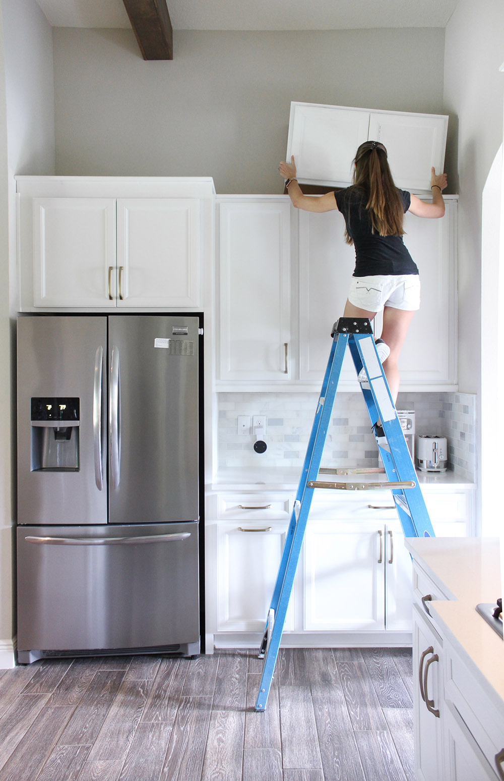 A woman standing on a ladder adds a smaller row of cabinets to the top of the current kitchen cabinets.