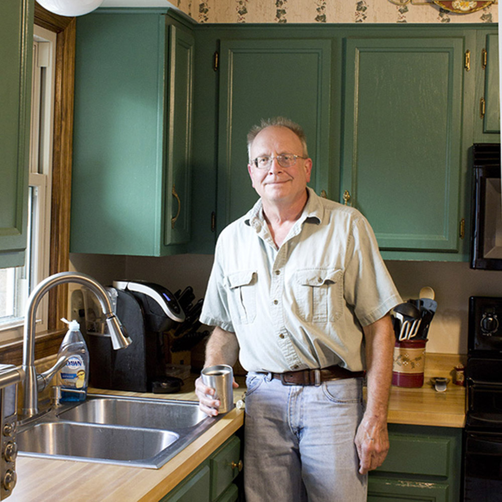 A man standing in a kitchen with green cabinets and wood countertops.