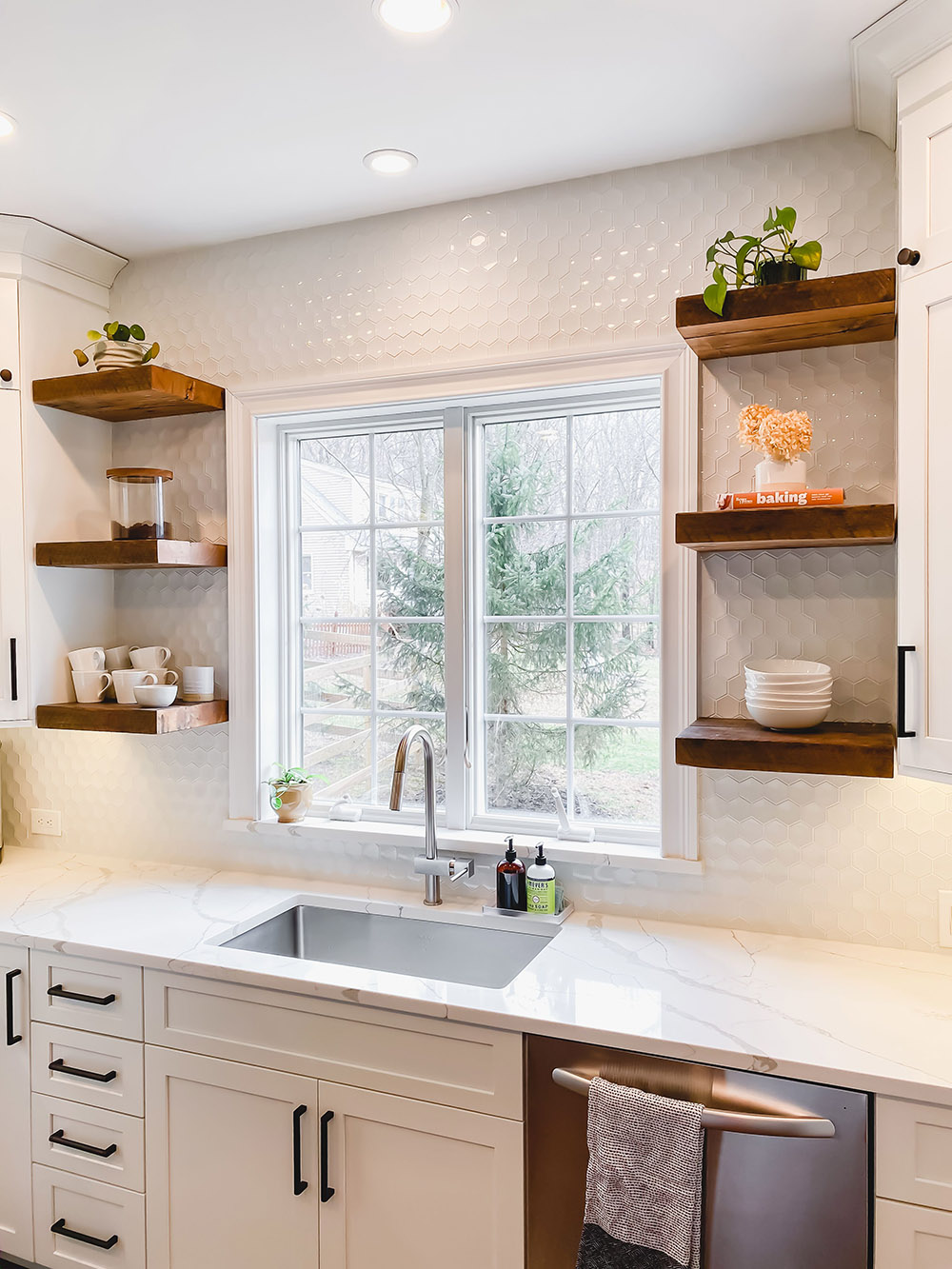 A kitchen sink area with a window surrounded by a tile wall with floating wood shelves.