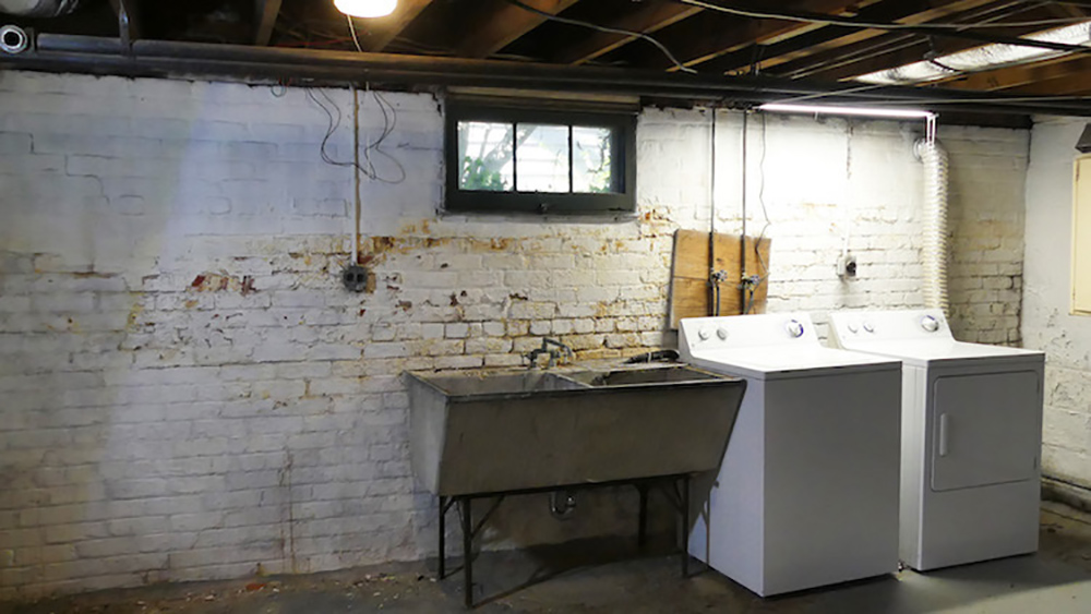 An old white washer and dryer sit next to a metal sink in front of a dirty white brick wall.