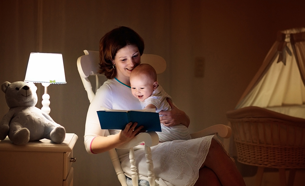 A mother sits with a baby in a room lit by a table lamp.