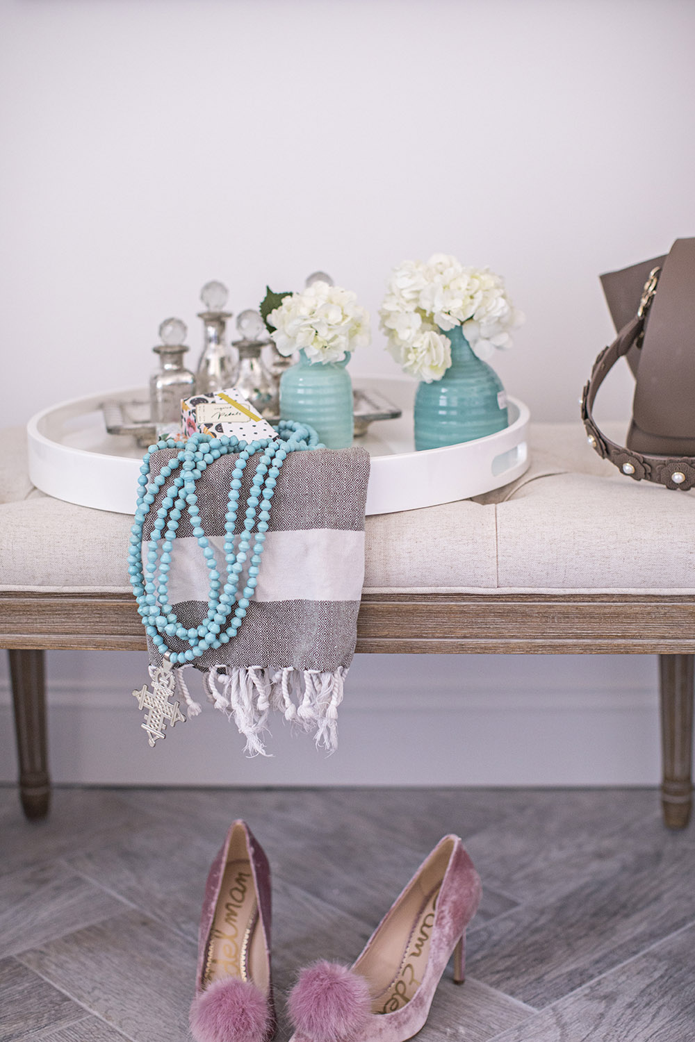 A white tray decorated with a throw and teal vases with flowers sitting on a bench.