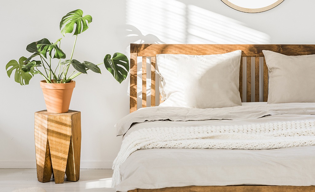 A Monstera plant on a wooden plant stand getting sunlight near a bed in a bedroom.