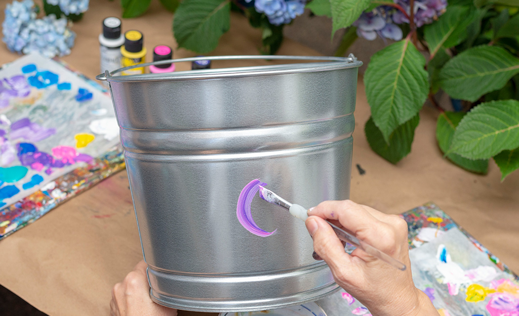 A person applying purple acrylic paint onto a galvanized bucket.