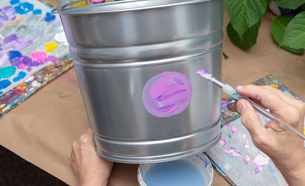 A person painting a purple circle onto a galvanized bucket.