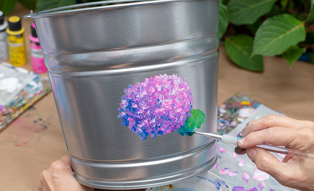 A person adding green paint to a galvanized bucket.