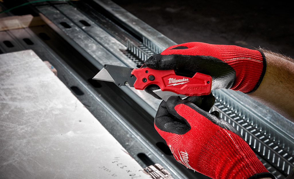 A person wearing red work gloves holds a utility knife.
