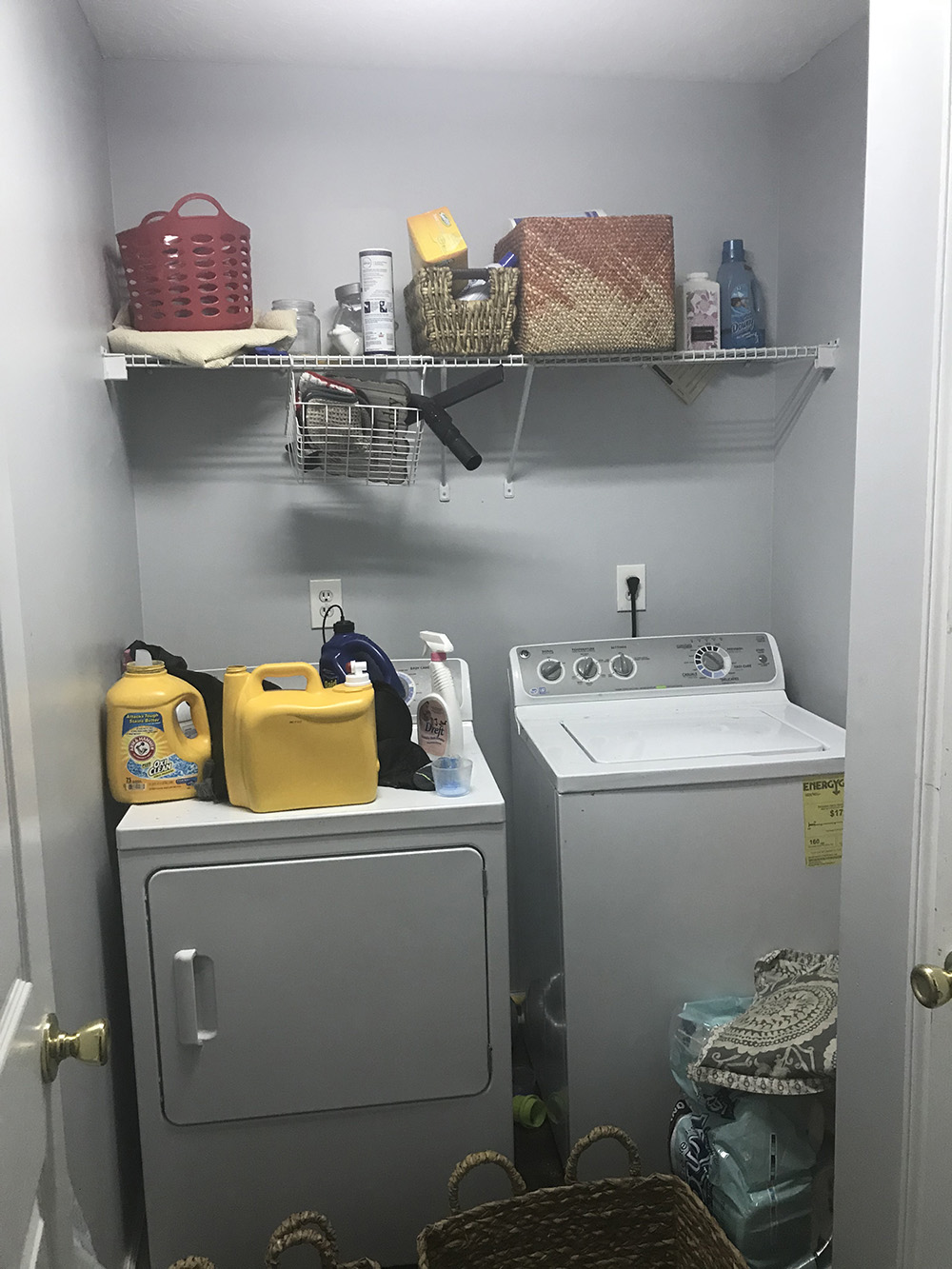 A small and unorganized laundry room with one wire shelf and white laundry appliances. 