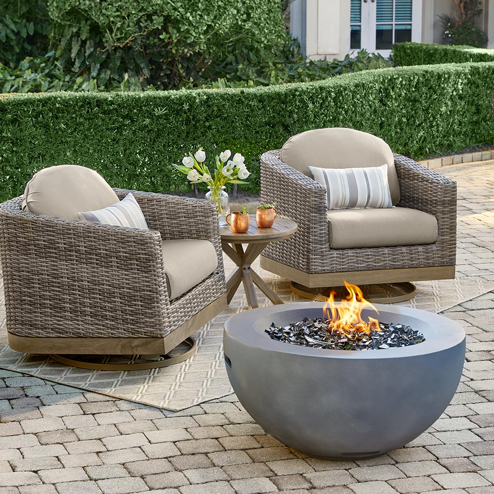 A bowl-shaped fire pit sits in front of two patio chair with beige cushions and a small side tale on a brick patio.
