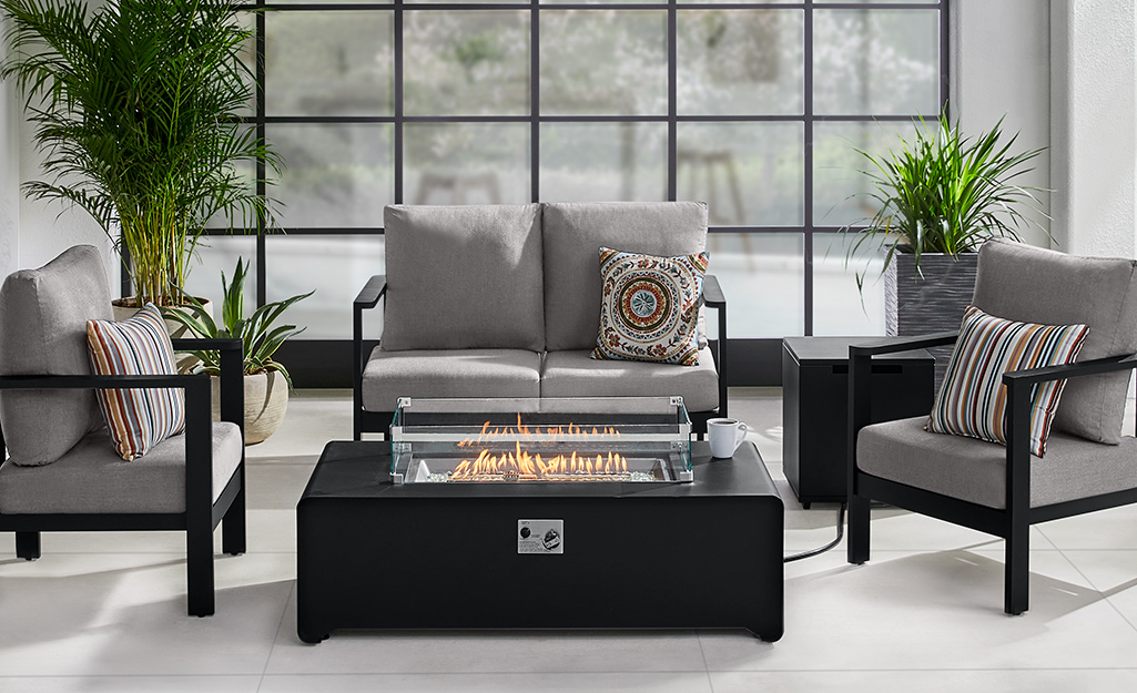 A fire pit table stands in the center of a patio conversation set with a love seat and two chairs, all with gray cushions.