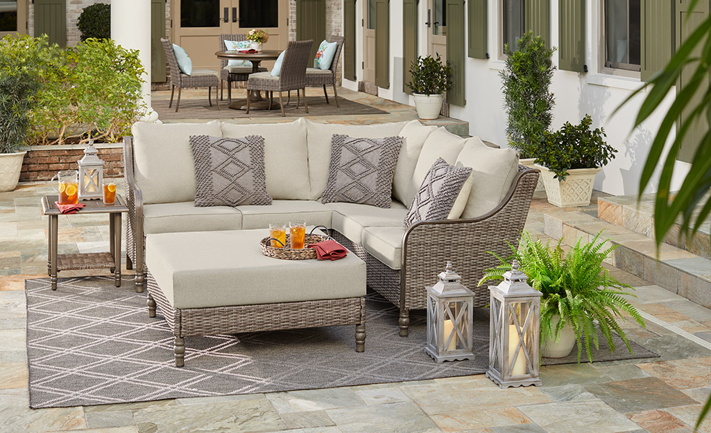 A neutral outdoor rug sits under a sectional sofa with beige cushions and matching ottoman on a large patio with an outdoor table and chairs in the background.