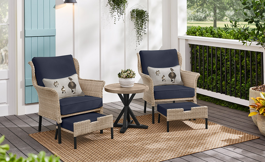 Two outdoors chairs with navy blue cushion and matching pull-out ottomans sit on a woven rug on a patio.