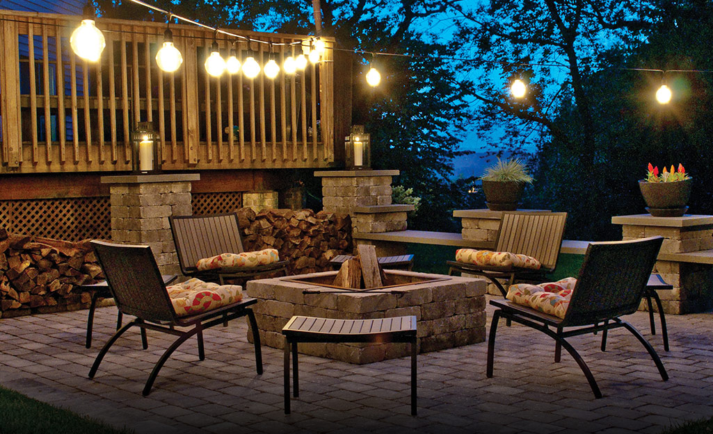 String lights illuminate a patio with a stone fire pit surrounded by four outdoor chairs with brightly patterned cushions.