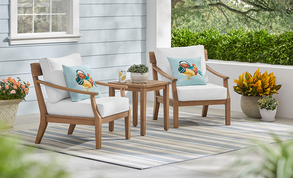 A side table sits between two wood chairs with white cushions and blue throw pillows sitting on a striped outdoor rug on a patio.