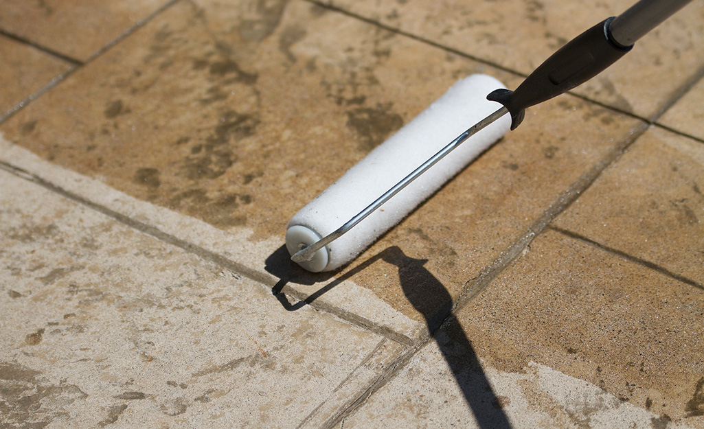 A person uses a paint roller to apply sealant to a patio surface.