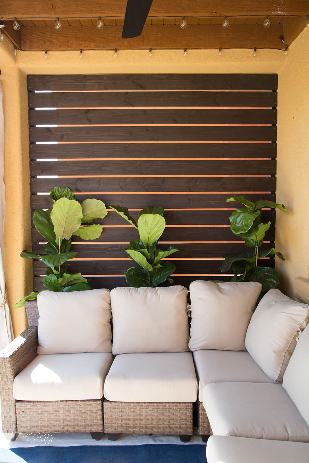 Planks of wood creating an outdoor wall with a beige patio couch and three plants.