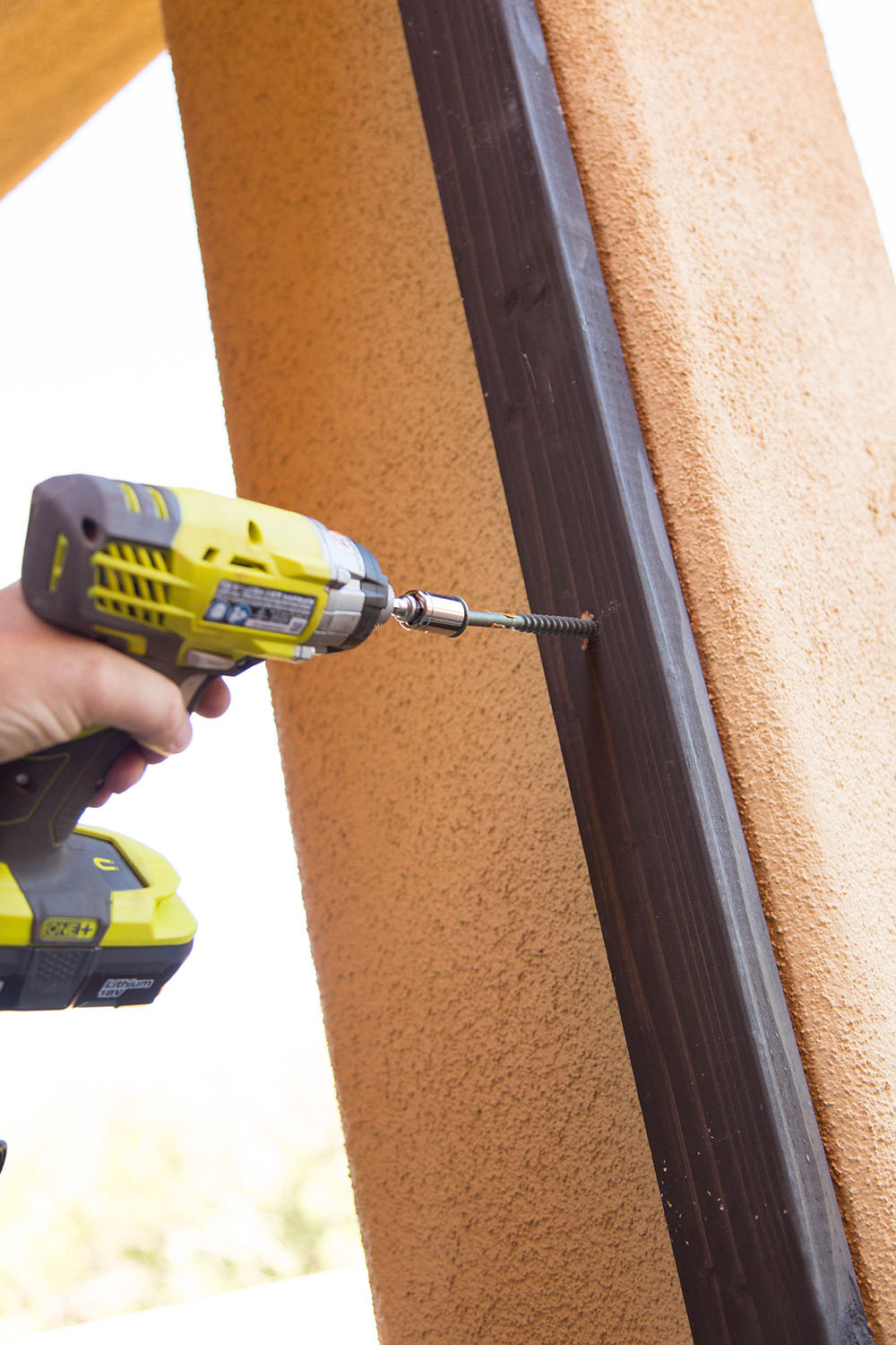 A man drilling a hole into wood attached to an outdoor wall.