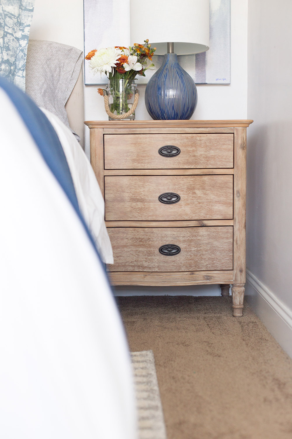 A vase of flowers and a blue lamp decorate a nightstand with three drawers.