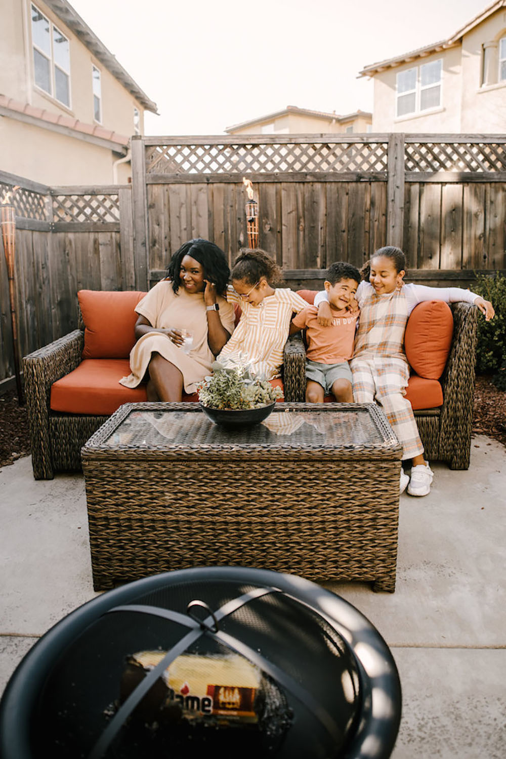 A woman and three children sit on a wicker patio set with bright cushions.