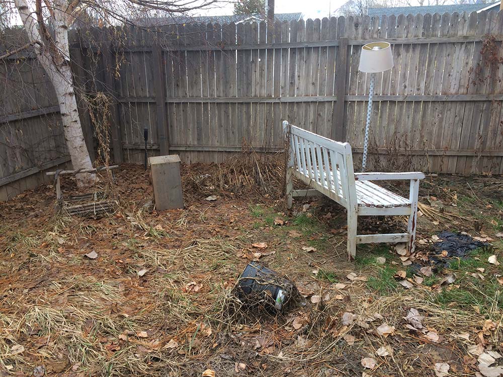 A fenced in backyard with an old lamp, bench, and overgrown weeds.