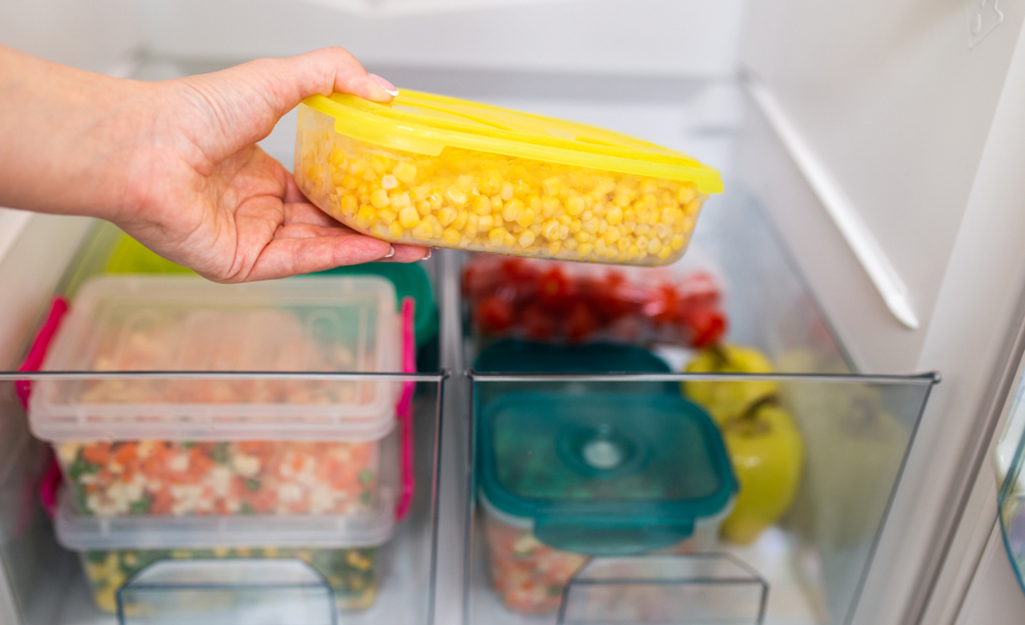 A person places a container of leftover corn in a refrigerator drawer.