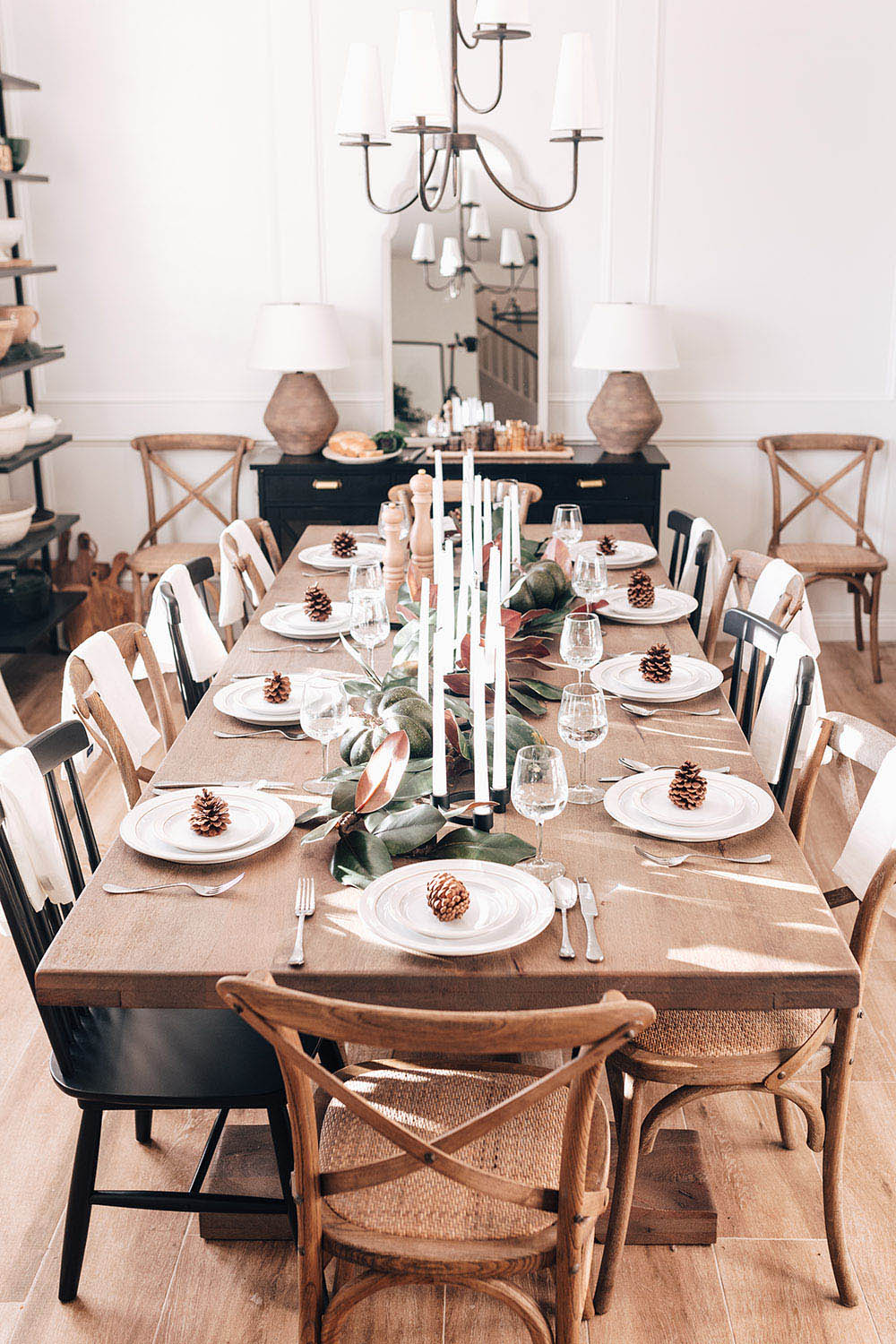 A rustic tablescape with white plates, pine cones, candles, and greenery.