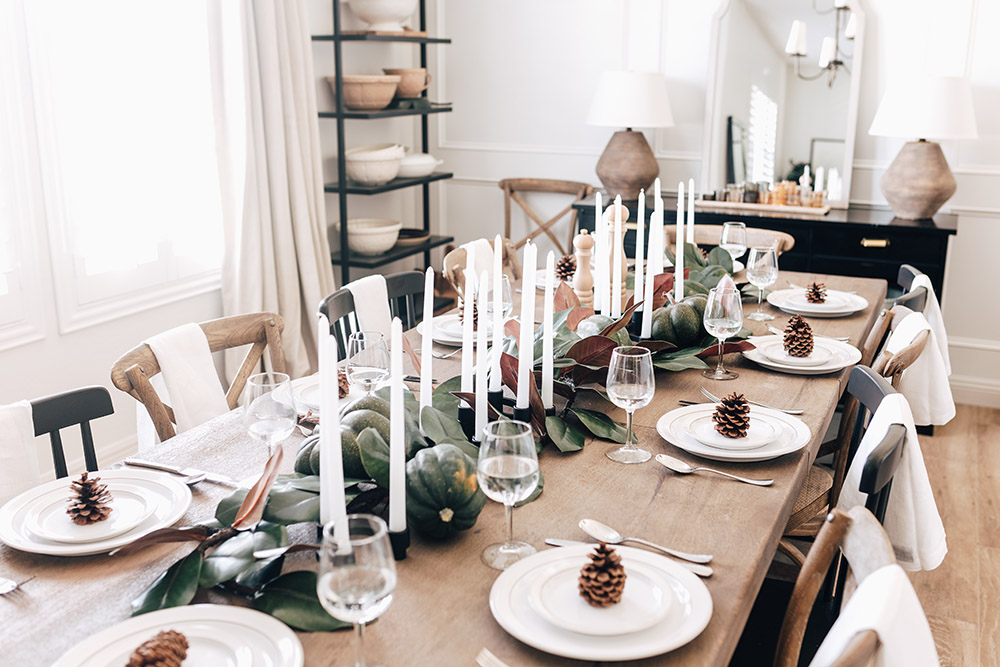 A rustic tablescape with white plates, pine cones, candles, and greenery.