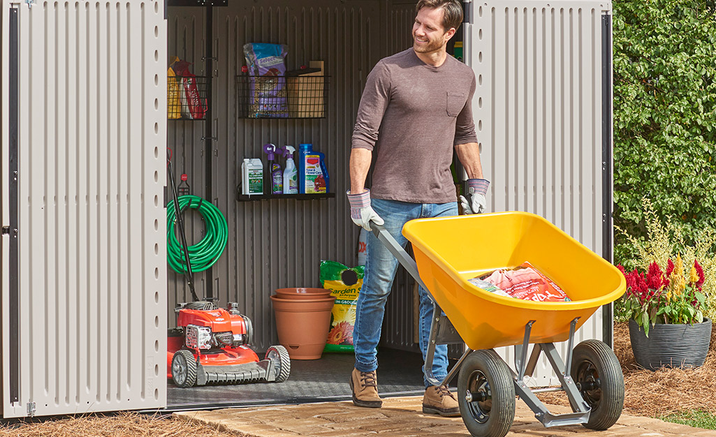 A person uses a wheelbarrow to take a bag of potting soil from a shed.