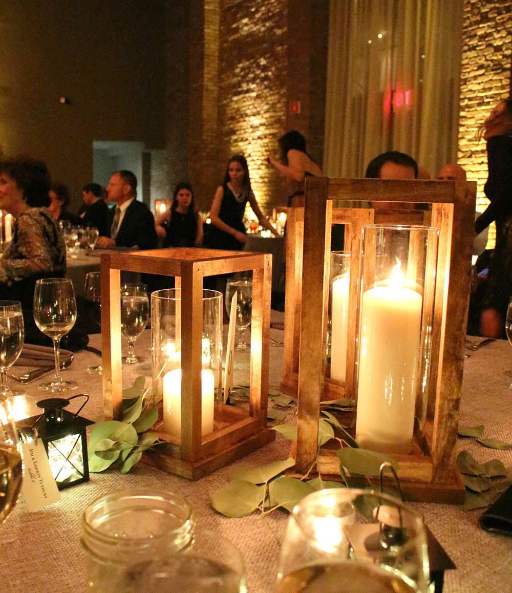 A dimly lit wedding reception with people behind a table of DIY wooden lanterns filled by candlelight.