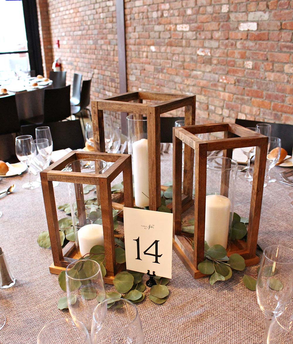 A table decorated with DIY wooden lanterns and eucalyptus leaves in front of a brick wall.