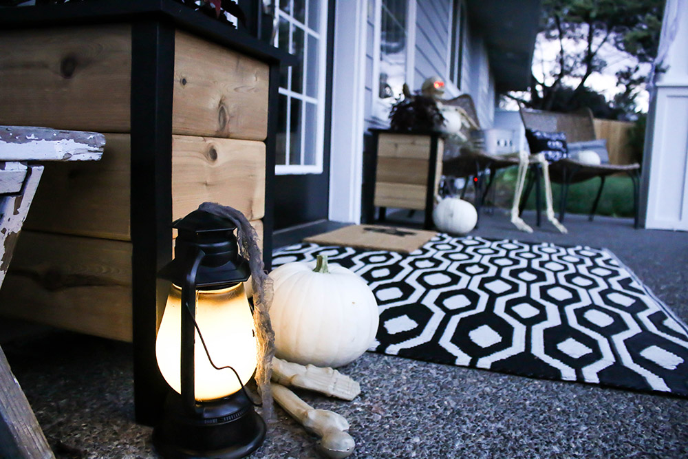 A front porch floor decorated with a white and black geometric rug, white pumpkins, and black lanterns.