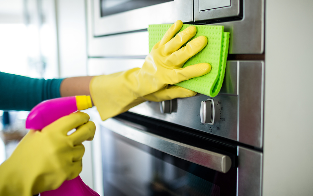 A person wearing gloves and cleaning the exterior of a wall oven with a sponge and spray bottle