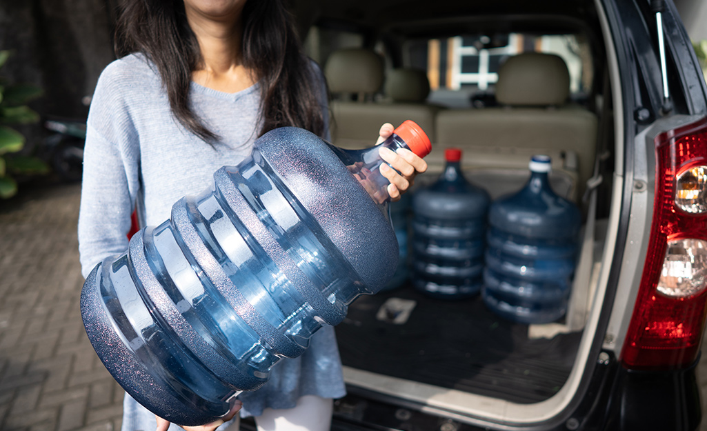 A woman puts a large water jug into the trunk of her car.