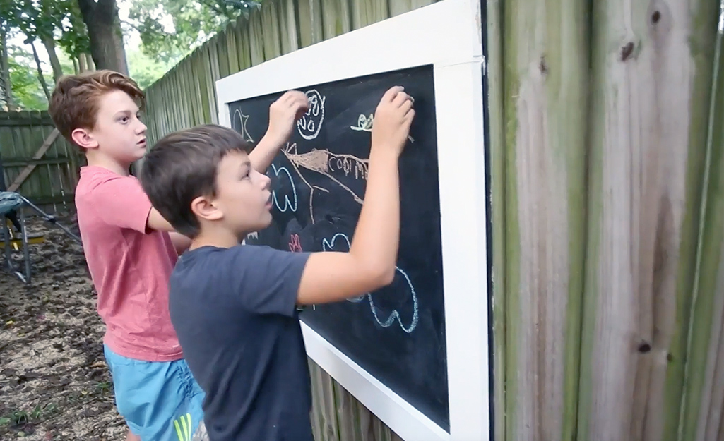 Two kids drawing on an outdoor chalkboard. 