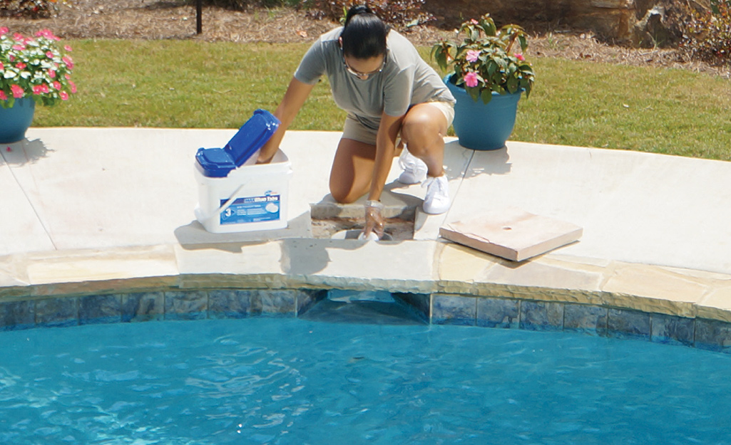 A woman kneels next to a swimming pool as she adds treatment to it.