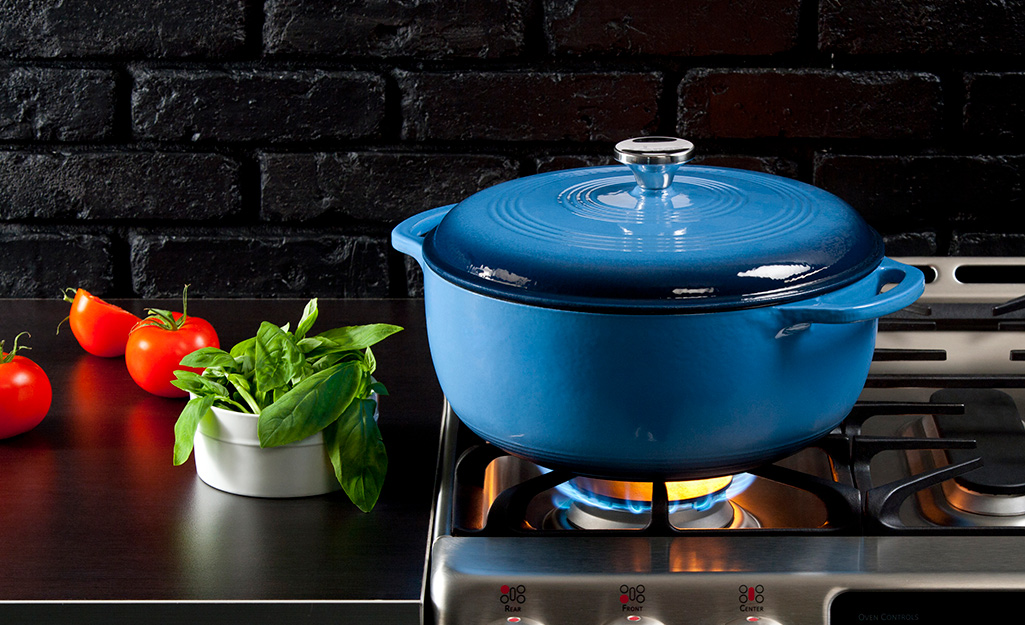 A blue Dutch oven on a stove top with a flame under it and herbs on the counter.