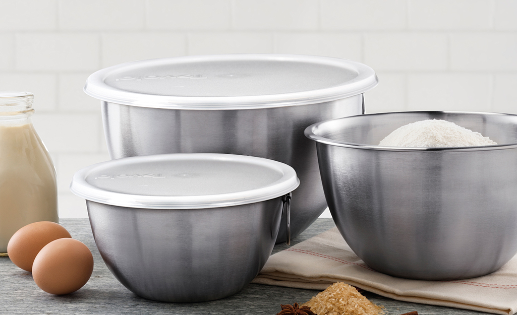 Three stainless steel mixing bowls, two with lids, stand on a counter.
