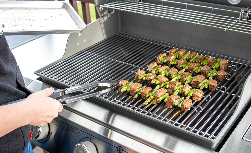 A person uses grilling tongs to rotate kebabs on a gas grill.